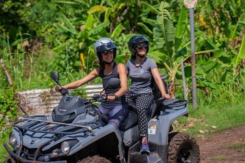 Two people on a quad bike in a rural setting with greenery and a dirt path. 5