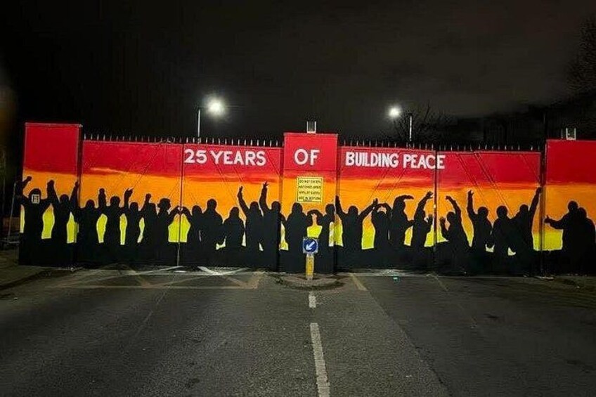 Une foule nombreuse se détache sur un mur rouge et jaune, sur lequel on peut lire l’inscription « 25 ANS AU SERVICE DE LA PAIX », et un panneau indicateur est visible au premier plan. 2