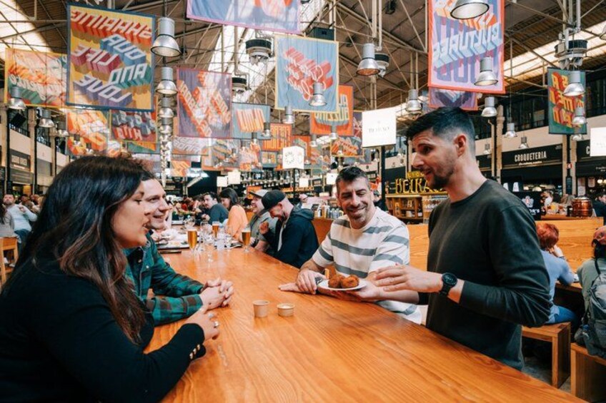 A bustling indoor market with wooden tables, people dining, and colorful banners. 1