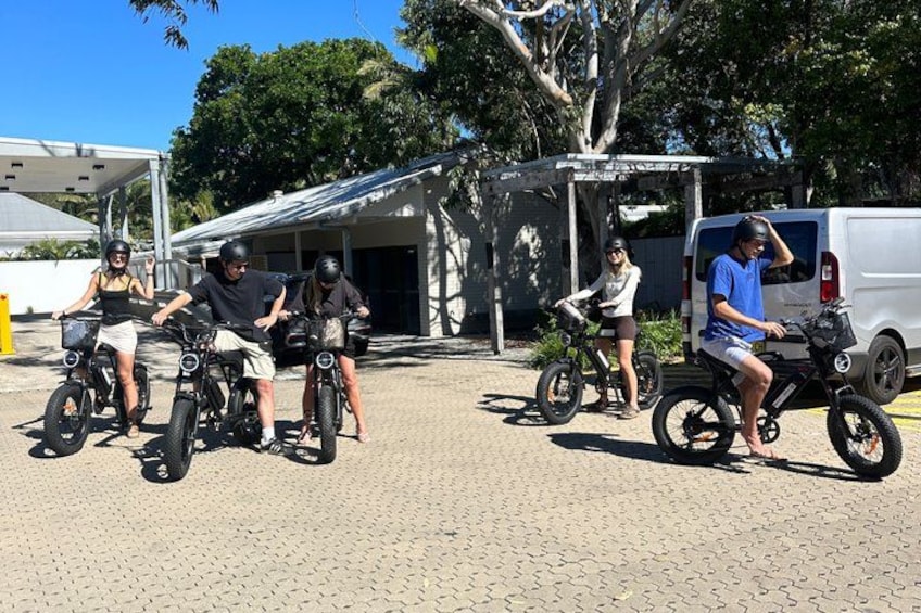 People riding electric bikes, a white van, and a covered structure. 1