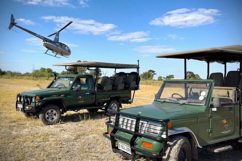 A green safari vehicle with a roof rack and a canopy, a helicopter in the background, and a dry grassland landscape. 3