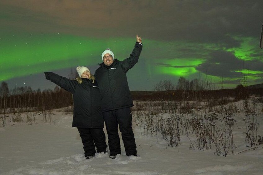 Two people standing in a snowy field under a green aurora. 2