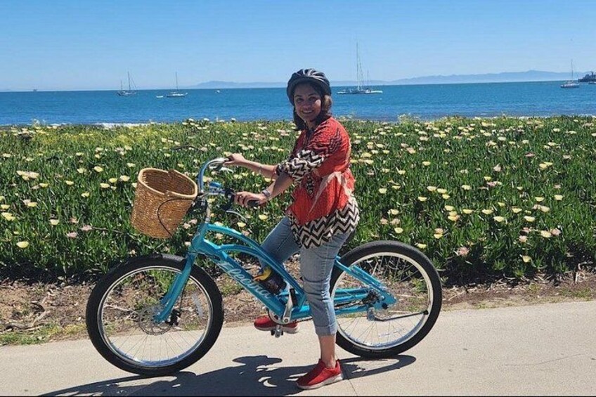 A person on a blue bicycle with a basket, standing on a paved path with a field of yellow flowers and a body of water in the background. 2