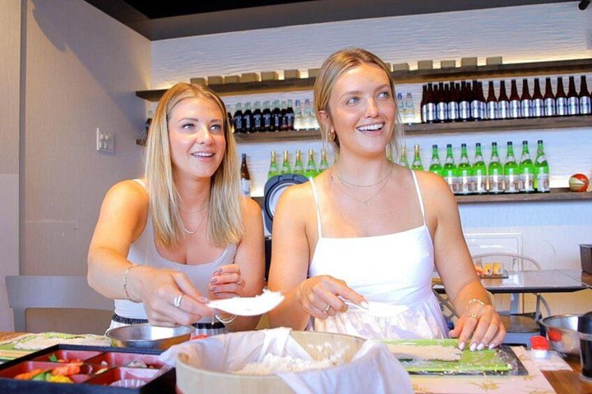 Two people preparing food with vegetables and a bottle of sauce on a table. 2