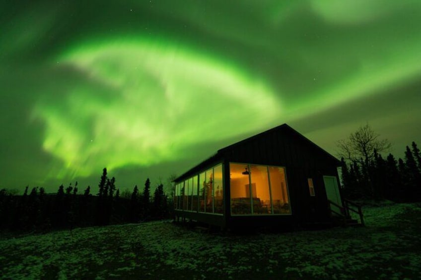 A glass-roofed cabin with a wooden exterior, surrounded by snow-covered ground and trees, under a green aurora display. 5