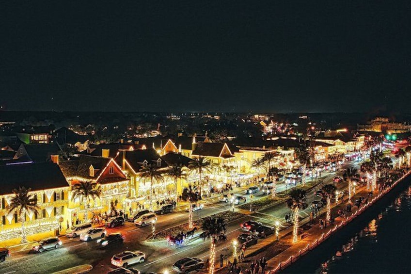 A brightly lit coastal town at night with illuminated buildings, a busy street, and palm trees lining the road. 2