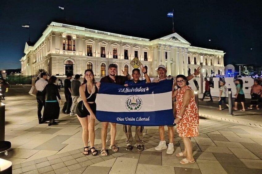 Un grupo de personas sostiene una bandera frente a un amplio edificio blanco con columnas. 1