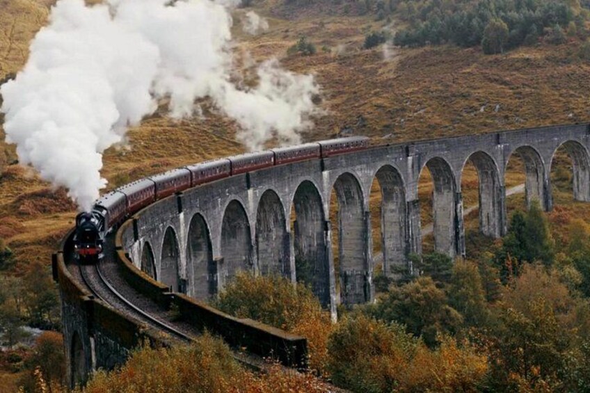Una locomotora de vapor sobre un puente ferroviario curvo, rodeada de árboles otoñales. 1