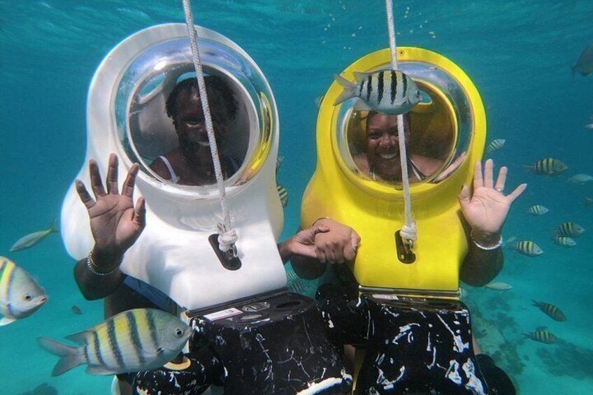 Two people wearing large, round, transparent helmets with ropes, underwater with fish swimming around. 2