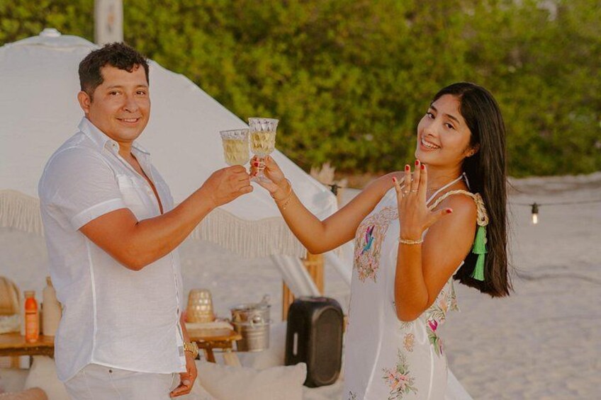 A couple toasting with drinks on a beach with a tent and greenery in the background. 2