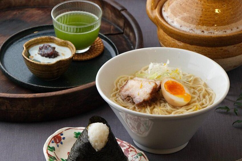 Een kom ramen met varkensvlees en een half gekookt ei, een klein schaaltje ingelegde pruim, een groen drankje, en een bord met een sushi rol. 1