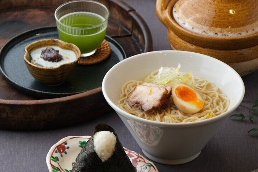 Un bol de ramen au porc, avec un œuf mollet, un petit bol de prunes marinées, une boisson verte et un morceau de sushi, le tout sur une table. 1