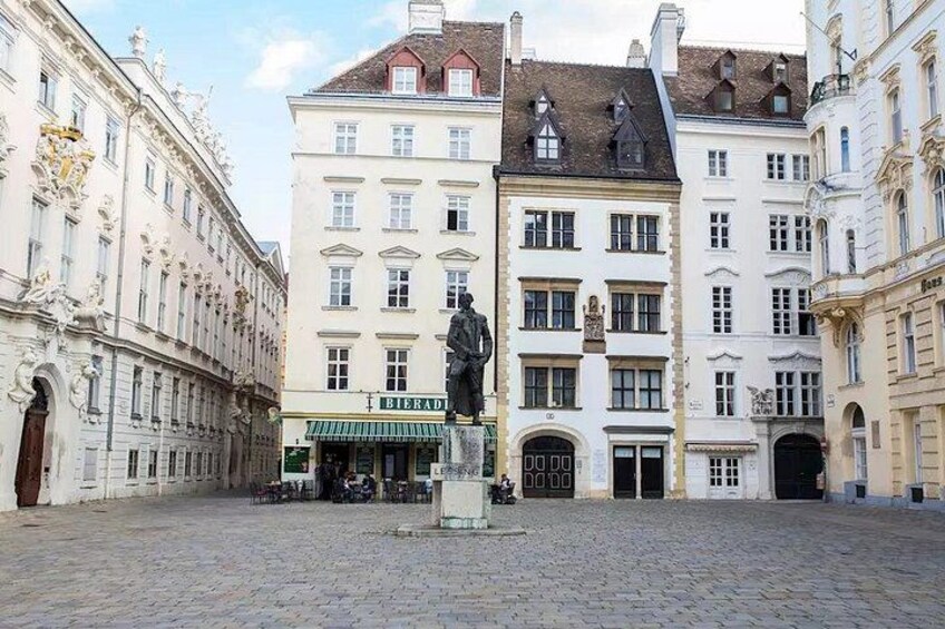 En brostensbelagt plads med historiske bygninger, en statue og et skilt for øl. 5
