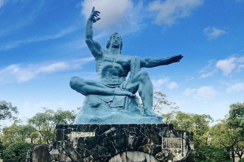 Una statua in bronzo raffigura una figura seduta con un braccio alzato, posata su un piedistallo di pietra con iscrizioni, circondata da alberi e un cielo sereno. 4