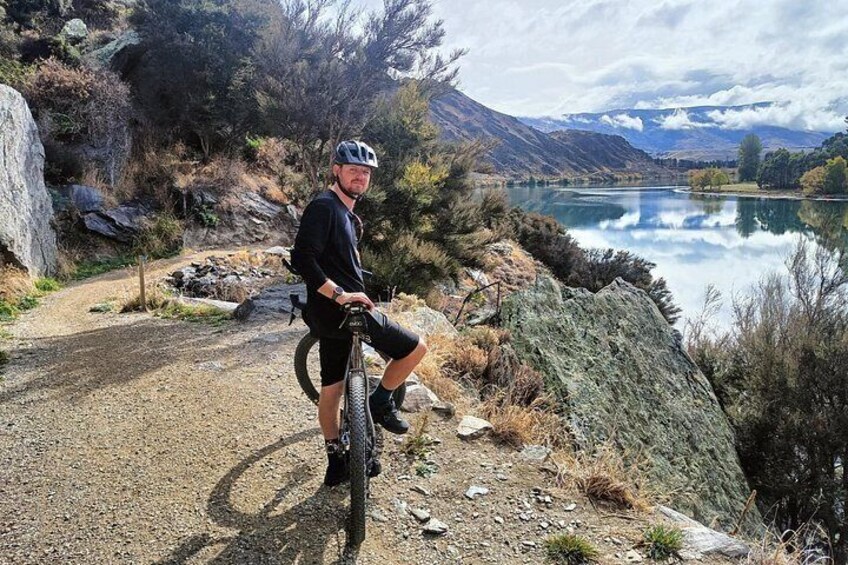 A person on a mountain bike on a gravel path near a lake with mountains in the background. 2
