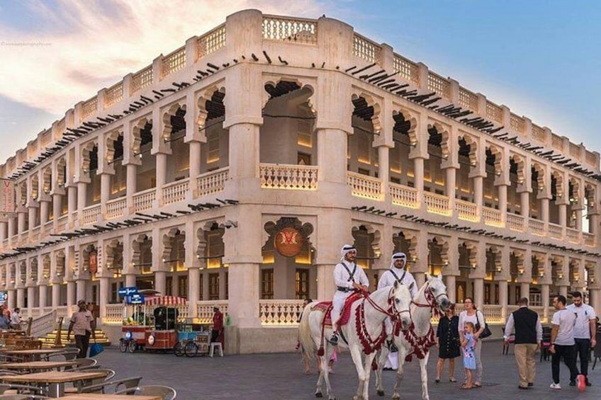 A historic building with multiple arches and columns, a clock, and people walking and riding horses. 4