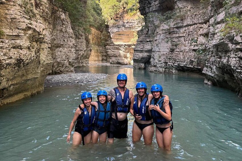 A group of people in blue helmets and life vests standing in a river surrounded by rocky cliffs. 5