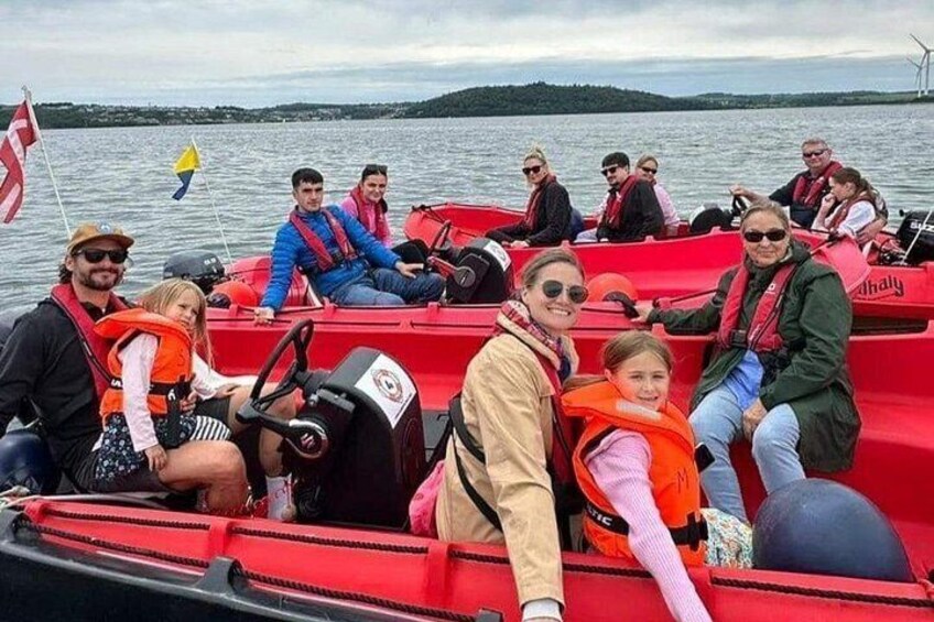 Un groupe de personnes se trouve sur un bateau rouge, portant des gilets de sauvetage. On aperçoit un lac et des éoliennes en arrière-plan. 2