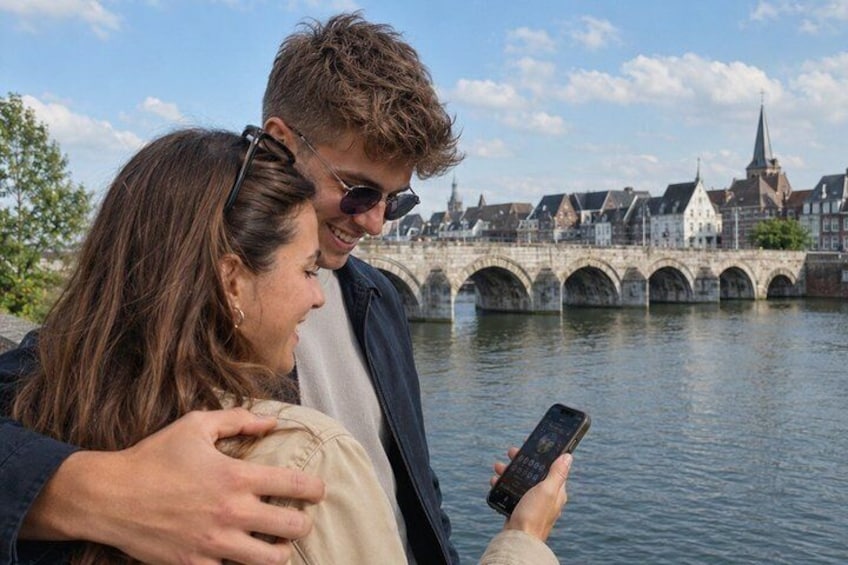 A couple standing by a river with a historic bridge in the background. 1