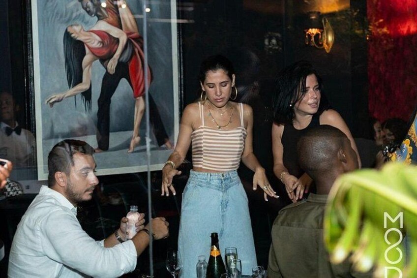 Escena de bar con gente socializando, un cartel de baile en la pared y botellas en la barra. 4