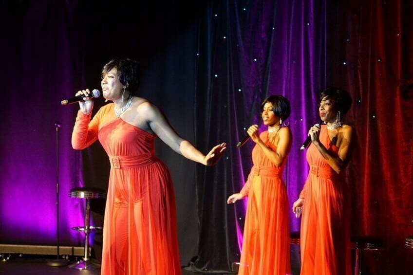 A stage with three performers in orange dresses, a microphone stand, and a drum set in the background. 2