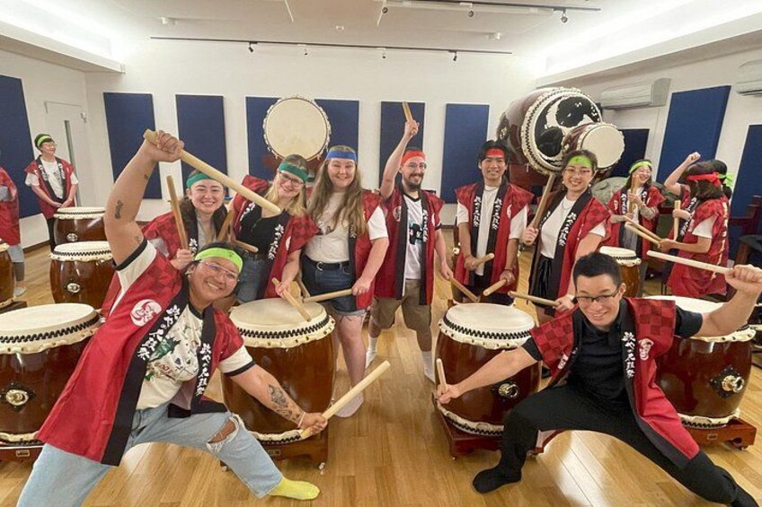 A group of people in a room with large drums, wearing red and black uniforms, and some with headbands. 1