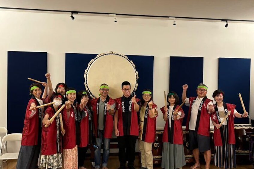 A group of people in red and black uniforms holding drums, standing in front of a large drum. 3