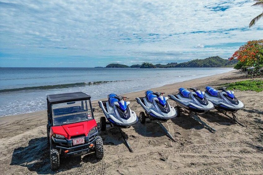 A red off-road vehicle and three jet skis on a sandy beach with the ocean in the background. 3