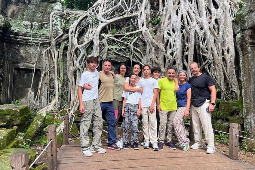 A group of people standing on a wooden platform with a backdrop of ancient stone structures and overgrown roots. 2