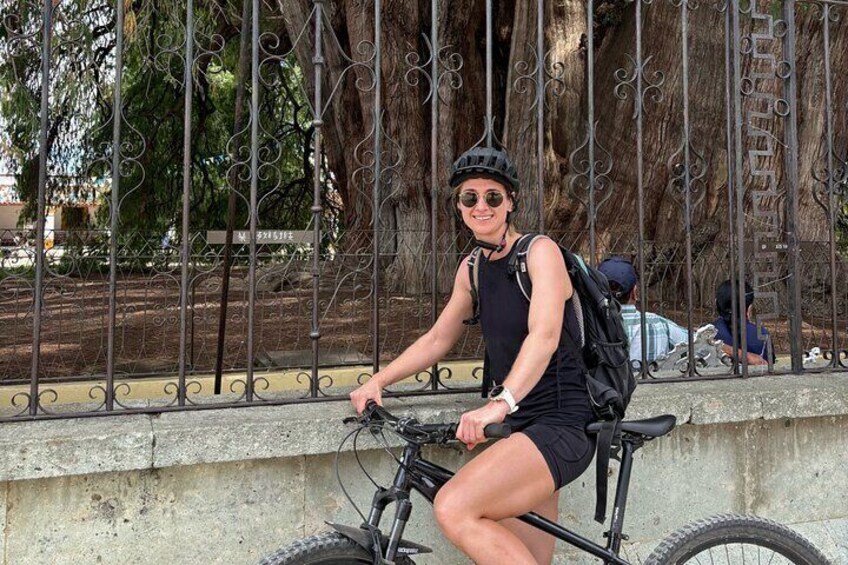 A person riding a bicycle with a backpack, wearing a helmet and sunglasses, in an outdoor setting with a metal fence and trees in the background. 2