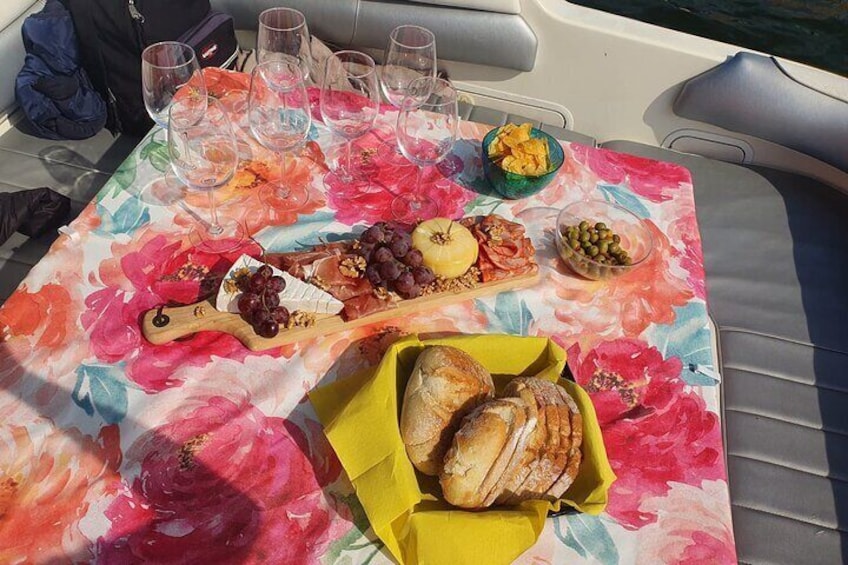 Une table recouverte d’une nappe à motifs floraux, une planche en bois servant de plateau avec des raisins et du fromage, un bol d’olives, une assiette de pain et plusieurs verres. 2