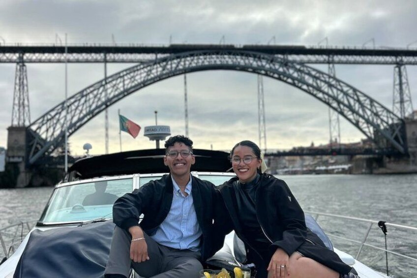 Un bote con una persona vestida de negro y otra con camisa azul, un puente al fondo y una bandera en el bote. 3