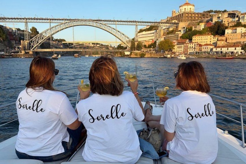 Tre persone su una barca, con la scritta 'Sorelle' sulle magliette, un ponte e uno skyline sullo sfondo. 1