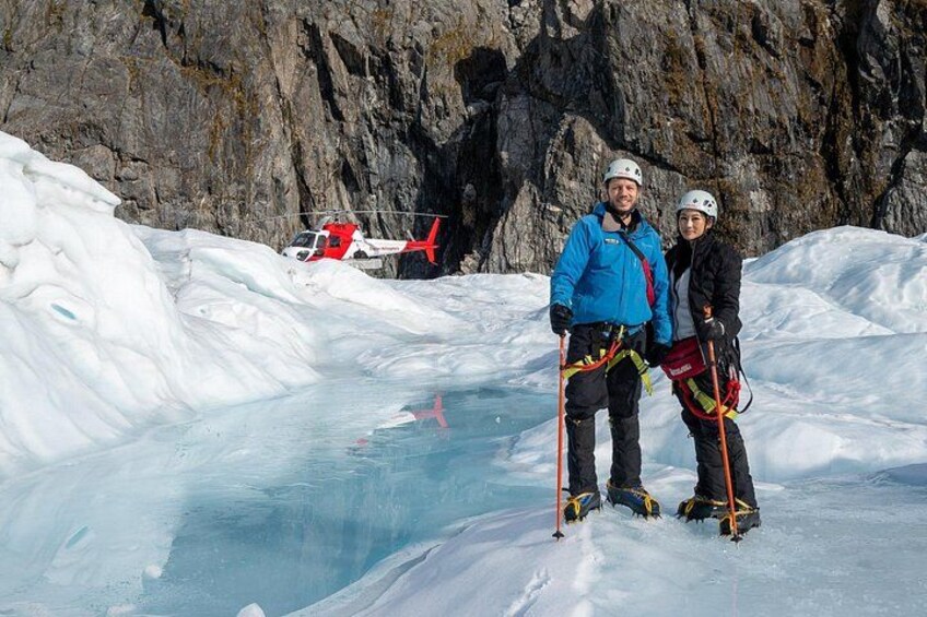 Two people with helmets and climbing gear on a snowy landscape with a helicopter in the background. 2