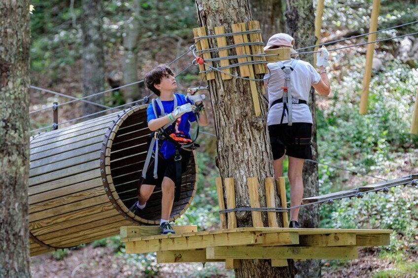 Dos personas en una aventura en un circuito boscoso, una escalando una estructura de madera y la otra ayudando. 2