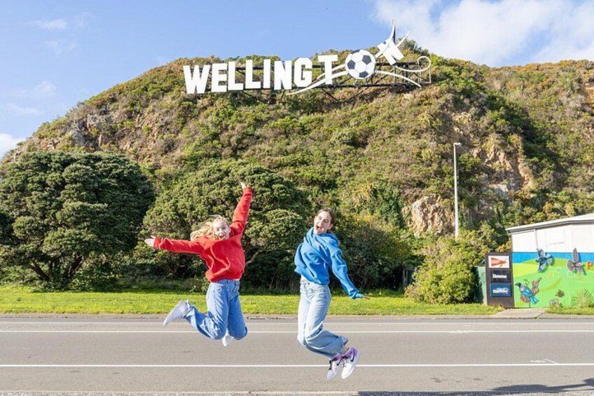 Two people jumping in front of a sign with 'WELLINGTOWN' and a soccer ball graphic. 3