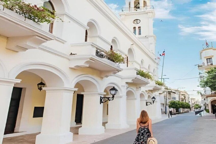 Una mujer caminando por una calle con edificios blancos, arcos y una torre del reloj. 1