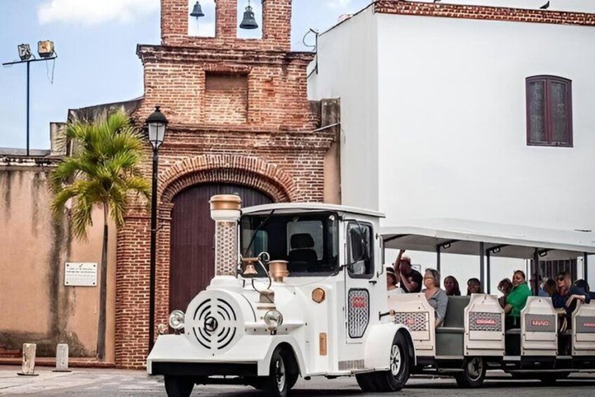 Un tranvía antiguo blanco con pasajeros, estacionado frente a un edificio de ladrillo con una torre de campanas. 2