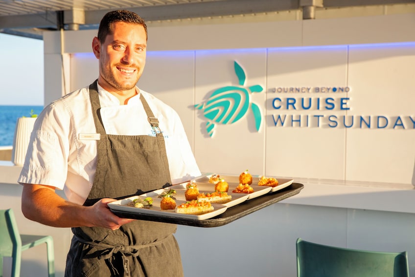 Henkilö kantaa tarjotinta, jossa on ruoka-annoksia, ja hän seisoo kyltin edessä, jossa lukee: ”Cruise Whitsunday”. 4