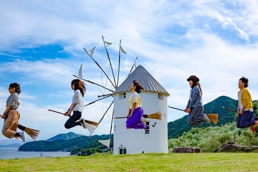 A group of people jumping in front of a windmill in a grassy area with mountains in the background. 4