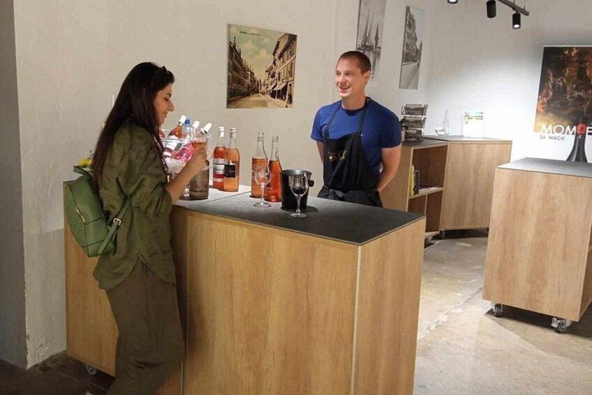 Una persona de pie junto a una barra de madera con una bolsa verde, un hombre con camisa azul y varias botellas y vasos sobre la barra. 2