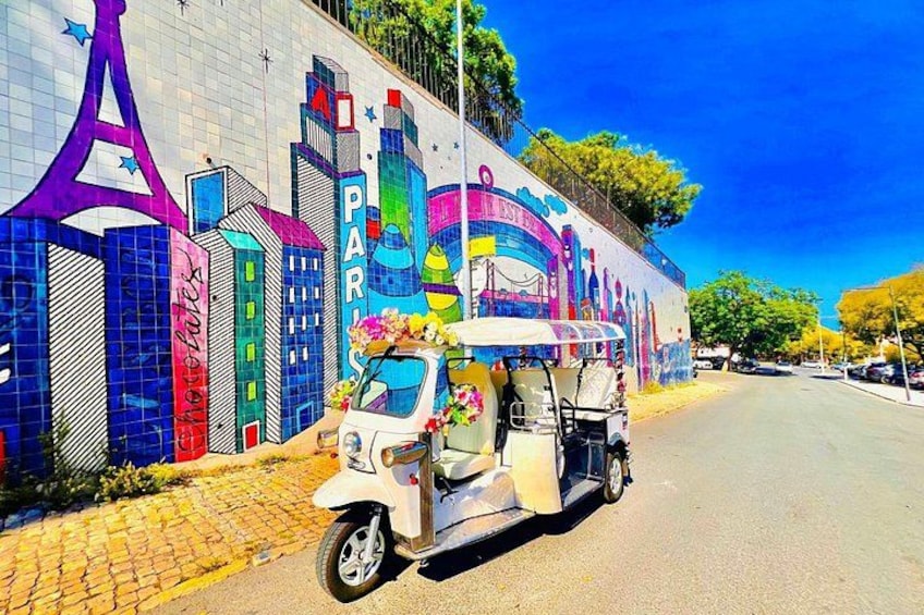 Un mural colorido con monumentos parisinos en una pared, un vehículo blanco de tres ruedas estacionado al frente, y un cielo azul despejado. 1