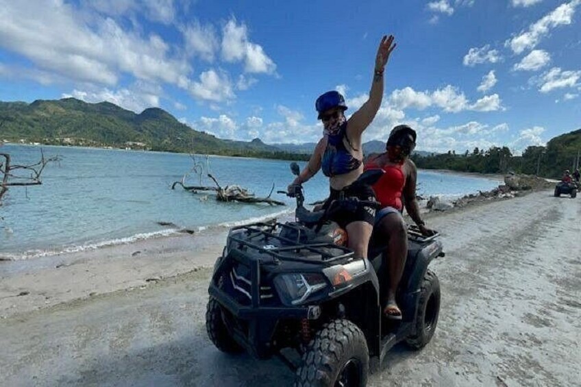 Dos personas en un cuatrimoto cerca de una playa, con agua y montañas al fondo. 3