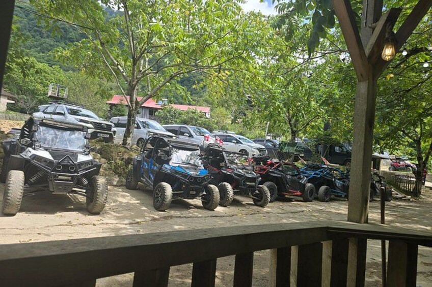 Off-road vehicles parked in a shaded area with trees and a wooden structure. 2