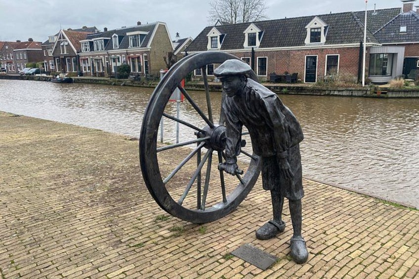 A statue of a man with a large wheel, a canal, and houses in the background. 3