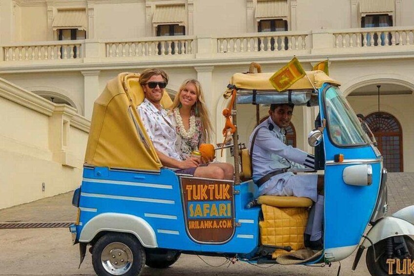 A tuk-tuk with a yellow canopy and blue body, labeled 'TUKTUK SAFARI,' parked in front of a building with balconies. 1