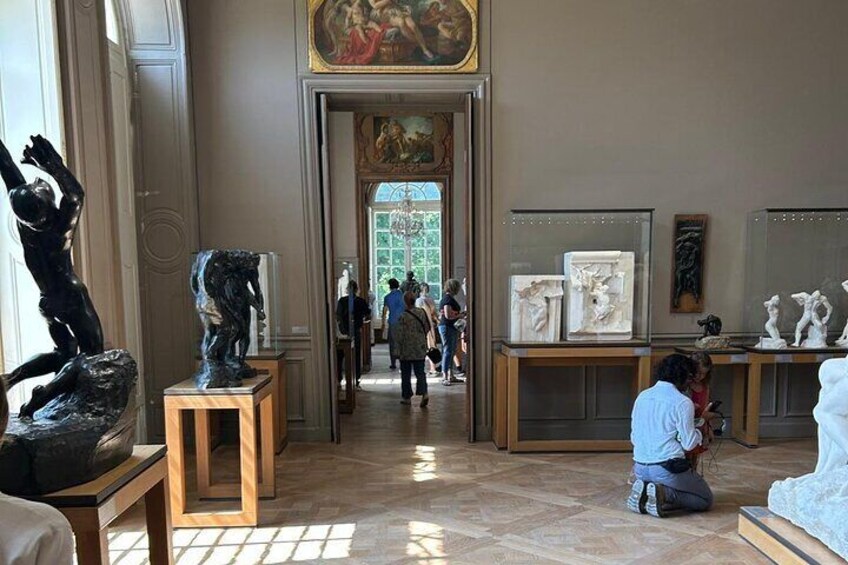 A museum interior with sculptures on pedestals, a central doorway, and visitors in the background. 1