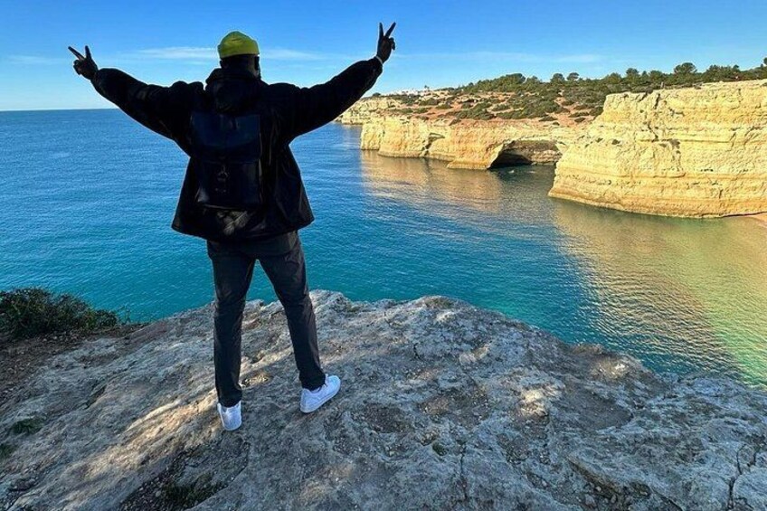 A person standing on a rocky cliff with arms outstretched, overlooking a coastal landscape with cliffs and a body of water. 1