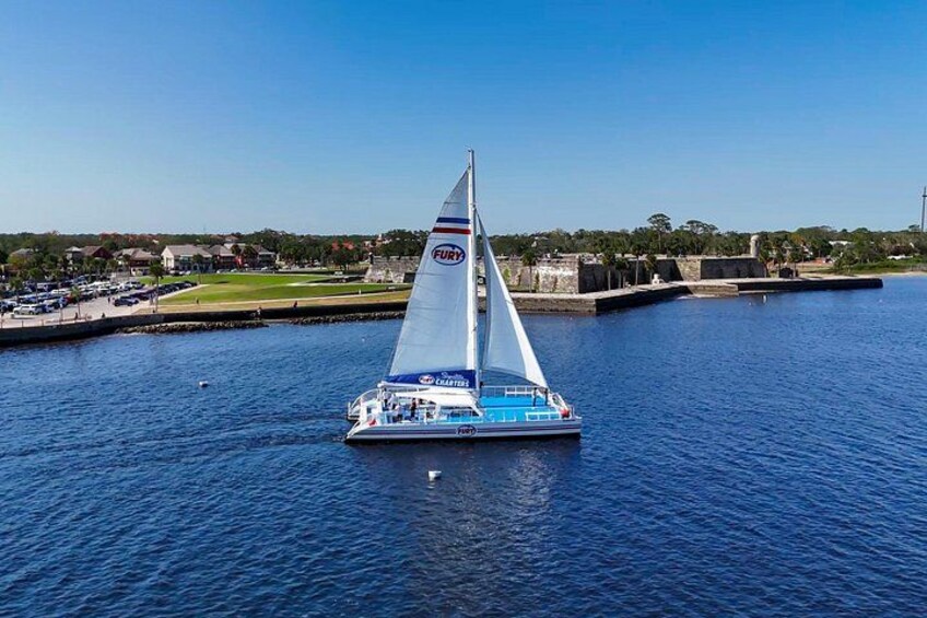 Een zeilboot met een blauw-wit onderwaterschip en een witte zeil, genaamd 'FURY', vaart over het water onder een heldere lucht, met een verre kustlijn met gebouwen en begroeiing. 5