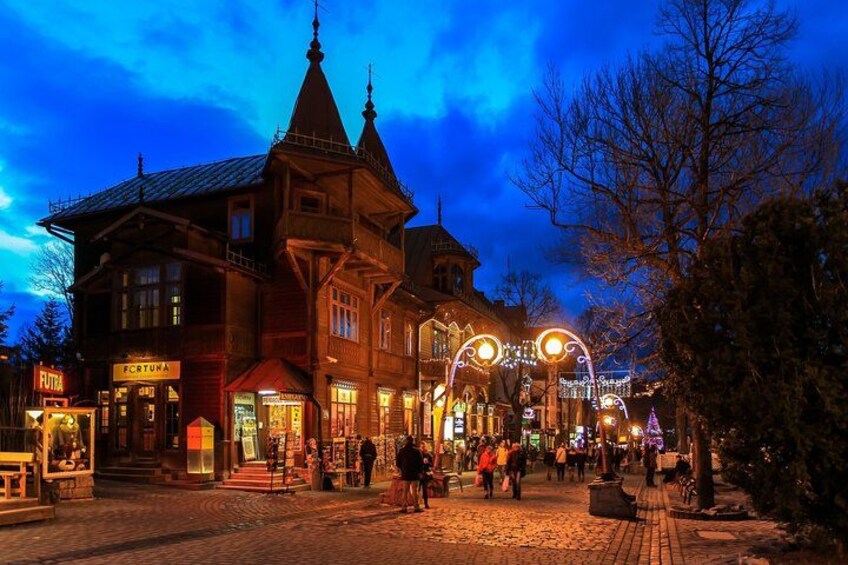 Een historisch gebouw met een puntig dak en een klokkentoren, omgeven door geplaveide straatjes en verlicht met feestelijke verlichting. 2